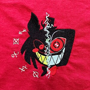 Op de afbeelding: Rood T-shirt met een geborduurd cartoonfiguur. Het ontwerp heeft een zwart silhouet, rode ogen en een brede grijns. De afbeelding is in tweeën gedeeld met witte stiksels. Geborduurde symbolen zijn ook aanwezig rond het personage.