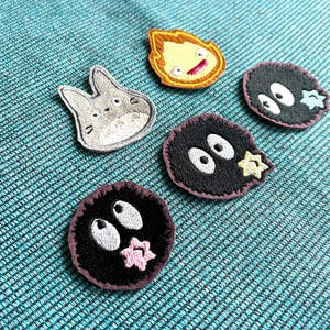 Op de afbeelding: Vijf geborduurde strijkpatches met personages uit de Studio Ghibli-film, Mijn Buurman Totoro. De patches zijn in verschillende kleuren en vormen, waaronder een grijs kat-achtig wezen, een gele vuurgeest en drie zwarte roetsprites.