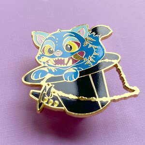 Op de afbeelding: Een blauwe kattenemaille pin met een zwarte hoge hoed. De kat heeft gele ogen en een grijnzende grijns. De pin heeft goudkleurige accenten en een gouden ketting. De achtergrond is lichtpaars.
