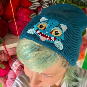 Peut inclure: Bonnet bleu sarcelle avec un visage de tigre de dessin animé brodé. Le tigre a des yeux jaunes, une langue rose et des dents blanches. Le bonnet est en tricot. L'arrière-plan montre de la laine colorée et des plantes.