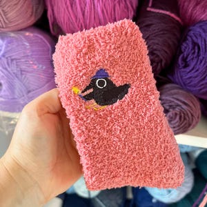 Può includere: Un calzino rosa soffice con un uccello nero ricamato che indossa un piccolo cappello viola. L'uccello è raffigurato con un becco giallo e zampe gialle, che risaltano sul materiale rosa morbido e testurizzato del tessuto del calzino.