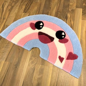 Puede incluir: Una alfombra semicircular con diseño de arcoíris. La alfombra tiene un borde azul claro, con rayas rosas, blancas y rosas. La alfombra presenta una cara sonriente con ojos negros y boca roja, y dos corazones rojos.
