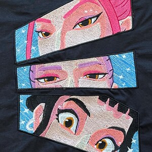 Könnte beinhalten: Gestickte Aufnäher mit stilisierten Anime-Augen mit rosa, lila und schwarzem Haar. Die Augen haben braune Iris und sind vor einem blauen Hintergrund mit weißen Sternakzenten platziert. Die Aufnäher befinden sich auf einem schwarzen Stoff.