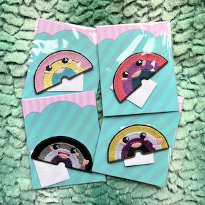 May include: Four embroidered iron-on patches featuring rainbow-shaped characters with different facial expressions. The patches are on white cardstock with a pink and blue striped background.