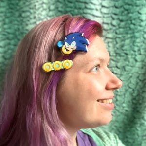 Peut inclure: Une femme avec des cheveux roses et violets porte deux pinces à cheveux. Une pince représente un dessin animé de Sonic the Hedgehog, et l'autre pince a trois anneaux jaunes et verts.
