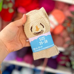 Op de afbeelding: Een paar beige sokken met een gebreid ruitpatroon en een geborduurd gezichtje. Een lichtblauwe papieren band zit om de sokken, met de tekst "SIZE 35-38" en "70% KATOEN, 27% POLYAMIDE & 3% ELASTANE" erop gedrukt.