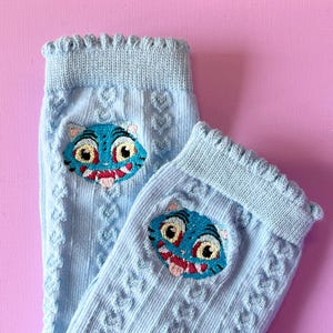 Puede incluir: Un par de calcetines azul claro con un patrón de corazones repetidos y un borde festoneado decorativo. Cada calcetín presenta una cara de tigre bordada en color turquesa con ojos amarillos, una lengua rosa y una boca roja.