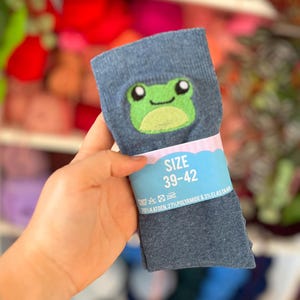 Op de afbeelding: Een paar blauw gemêleerde sokken met een geborduurd groen kikkergezicht op de boord. Een lichtblauwe papieren band om het midden toont de tekst SIZE 39-42, samen met wasvoorschriften en details over de materiaalsamenstelling van de sokken.