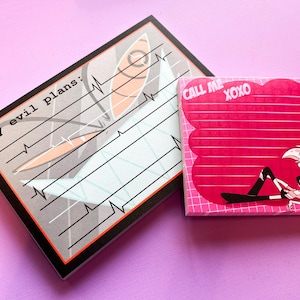 Puede incluir: Dos blocs de notas con gráficos de dibujos animados. Un bloc de notas tiene el texto "My evil plans:" con un diseño estilizado. El otro bloc de notas tiene el texto "CALL ME XOXO" y una ilustración de personaje de dibujos animados. Ambos blocs de notas están sobre una superficie púrpura.