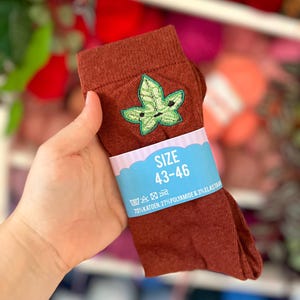 Op de afbeelding: Een paar roestkleurige sokken met een geborduurd groen bladmotief. Een lichtblauwe papieren band is om het midden gewikkeld, met de tekst SIZE 43-46, wassymbolen en de samenstelling 70% KATOEN, 27% POLYAMIDE & 3% ELASTAAN.