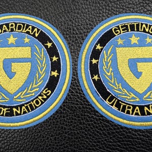 Può includere: Due toppe ricamate con sfondo blu, bordo nero e dettagli gialli. La toppa a sinistra dice "Guardian Units of Nations" e la toppa a destra dice "Getting Ultra Nasty".