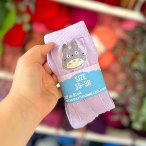 Op de afbeelding: Lichtpaarse sokken met een textuur van hartjes en een geborduurde grijze figuurpatch. Een blauwe papieren band zit om het midden met de tekst SIZE 35-38, wassymbolen en informatie over de materiaalsamenstelling van de sokken.