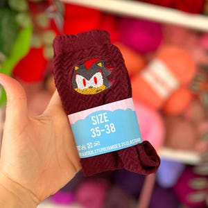 Op de afbeelding: Een paar donkerrode sokken met een textuur in visgraatmotief en een geborduurde afbeelding van het personage Shadow the Hedgehog. Een lichtblauwe papieren band zit om de sokken, met de tekst SIZE 35-38 en informatie over de materiaalsamenstelling.