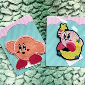May include: Two embroidered patches featuring a pink and yellow character with a star on its head. The character is holding a yellow crescent moon. The patches are on a pink and blue background.