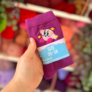 Op de afbeelding: Een paarse sok met een geborduurd roze figuurtje met blauwe ogen en gele sterren. Een lichtblauwe papieren band zit om het midden, met de tekst SIZE 35-38, wassymbolen en de samenstelling 70% KATOEN, 27% POLYAMIDE & 3% ELASTAAN.