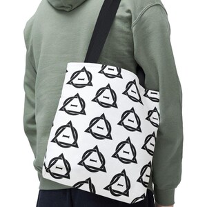 May include: White tote bag featuring a black geometric pattern of triangles and a horizontal line. The bag has a black strap and is worn over the shoulder. The background shows a person in a green hoodie.