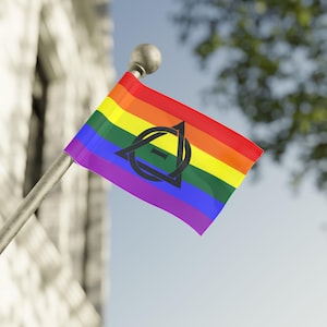 May include: A rainbow pride flag with a black symbol of a circle with a triangle inside, flying on a flagpole in front of a building.