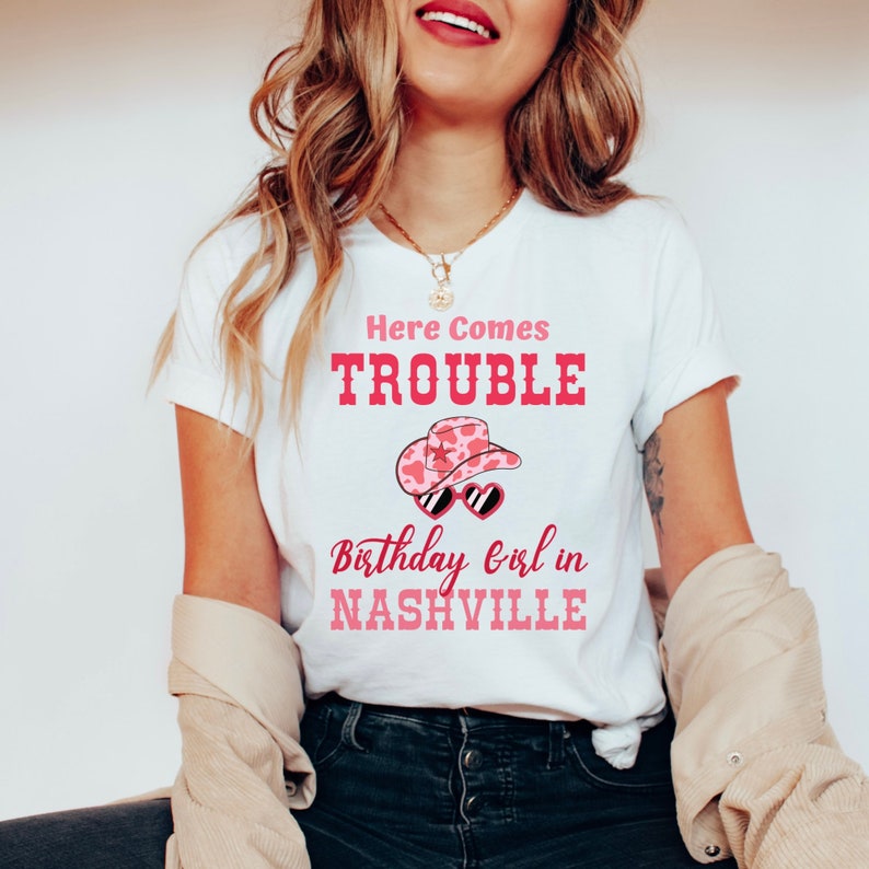 Pu&ograve; includere: T-shirt bianco con la scritta rosa "Here Comes Trouble". Sotto, un cappello da cowboy rosa con occhiali da sole e la scritta "Birthday Girl in Nashville".