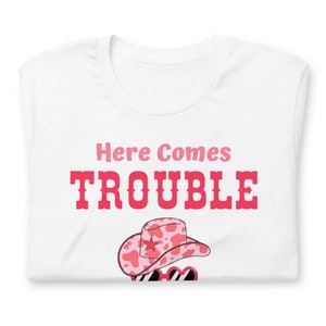 Pu&ograve; includere: T-shirt bianco con la scritta "Here Comes Trouble" in rosa e rosso. La maglietta presenta un cappello da cowboy rosa e bianco con stampa di mucca con una stella e occhiali da sole.