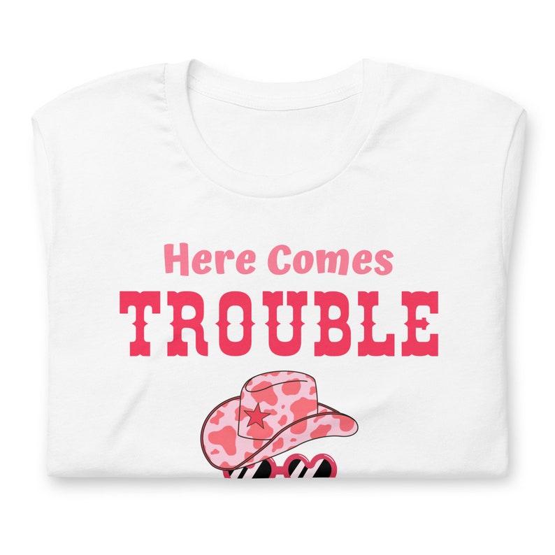 Pu&ograve; includere: T-shirt bianco con la scritta "Here Comes Trouble" in rosa e rosso. La maglietta presenta un cappello da cowboy rosa e bianco con stampa di mucca con una stella e occhiali da sole.