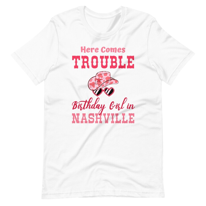 Pu&ograve; includere: T-shirt bianco con un design grafico rosa. Il design include il testo "Here Comes Trouble", un cappello da cowboy e occhiali da sole a forma di cuore, e il testo "Birthday girl in Nashville."