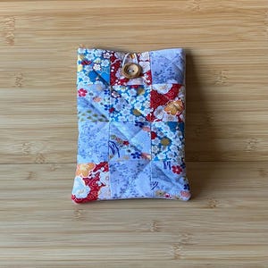 May include: A quilted fabric case with a patchwork design. The case features floral patterns in red, blue, and white. A wooden button and loop closure secures the top. The rectangular case is likely a sleeve for a small electronic device, such as a phone.