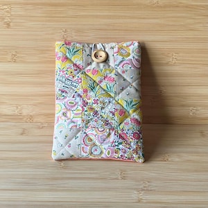 May include: A patchwork fabric case with a floral design. The case is made up of squares of fabric in yellow, pink, and white, with a wooden button fastening. The case is rectangular and likely for a phone or small tablet.
