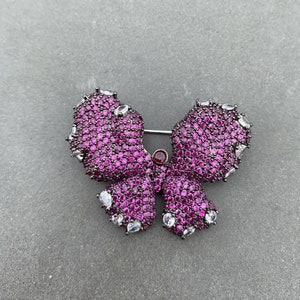 Könnte beinhalten: Eine rosa Schmetterlingsbrosche mit klaren und roten Strasssteinen. Die Brosche ist aus Metall und hat eine Nadel auf der Rückseite.