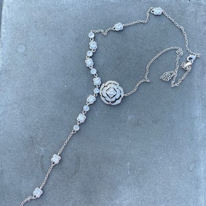 Pode incluir: Um colar de prata com um grande pingente de cristal transparente em forma de flor e pequenos detalhes de cristal transparente. O colar está sobre uma superfície cinza.