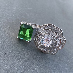May include: A silver ring featuring a large green emerald-cut gemstone and a flower design with smaller white gemstones.