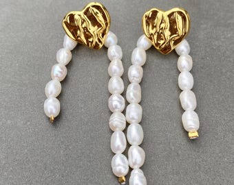 May include: A pair of gold-coloured heart-shaped earrings with a textured surface. Each earring features a strand of white, oval-shaped pearls. The earrings hang from the heart-shaped tops.