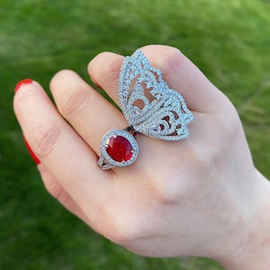 Puede incluir: Un anillo de plata con una gran piedra ovalada roja rodeada de piedras más pequeñas transparentes. El anillo se lleva en el mismo dedo que un anillo de mariposa de plata con piedras transparentes.