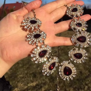 Puede incluir: Un collar de oro con un diseño grande y ornamentado que presenta múltiples piedras preciosas rojas en forma de lágrima rodeadas de diamantes de imitación transparentes.