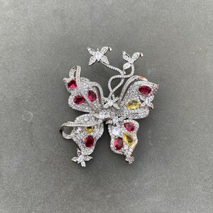 Könnte beinhalten: Eine silberne Schmetterlingsbrosche mit roten, gelben und klaren Edelsteinen. Der Schmetterling hat komplizierte Details und ist mit funkelnden Steinen besetzt.