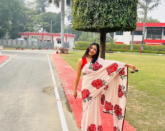 May include: A woman wearing a light pink saree with large red rose floral patterns. The saree has a dark red border. She is standing on a red brick path, with a green lawn and buildings in the background.