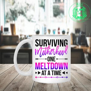 Könnte beinhalten: Ein weißer Keramikbecher mit dem schwarzen Text "SURVIVING", "ONE" und "AT A TIME", dem Wort "Motherhood" in einer violett-pinken Verlaufsschrift und "MELTDOWN" in violett-pinken Blockbuchstaben, verziert mit schwarzen Pfeilsymbolen.