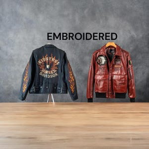 May include: Two embroidered leather jackets are displayed against a gray background. One is black with orange flame designs and Harley Davidson text. The other is a reddish-brown bomber jacket with patches and pockets. The word EMBROIDERED is above the jackets.