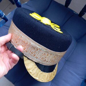Puede incluir: Un sombrero de fieltro negro con ribete dorado y un diseño bordado en amarillo. El sombrero tiene una ala ancha y una copa alta.
