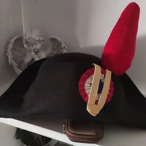 Puede incluir: Un sombrero bicornio negro con una pluma roja y una escarapela tricolor. El sombrero presenta una cinta dorada y un emblema circular con detalles rojos, blancos y azules. Es un estilo histórico.