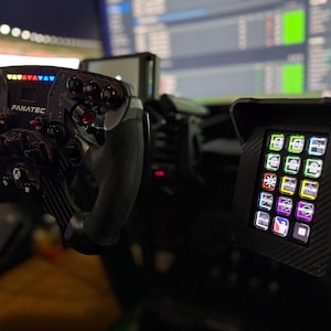 May include: A black and gray Fanatec racing wheel with multiple buttons and a display. A black control panel with illuminated buttons is to the right. The setup is for a racing simulator.
