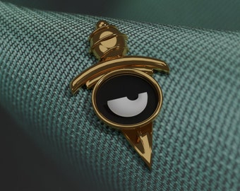 Puede incluir: Un pin de color dorado en forma de daga con un círculo negro y un diseño de media luna blanca.