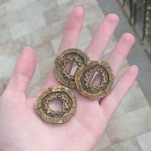 May include: Three gold-colored metal rings with intricate designs. Each ring has a circular opening in the center. The rings are arranged in a loose pile.