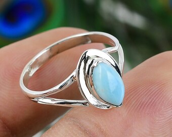 May include: A silver ring with a light blue, teardrop-shaped gemstone. The ring has a split shank design, with the gemstone held in a bezel setting. The ring is shown against a blurred background of green and blue.