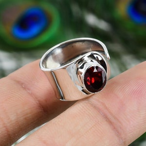 Puede incluir: Un anillo de plata con una piedra preciosa roja engastada en un engaste de bisel. El anillo es ajustable y tiene una banda ancha.