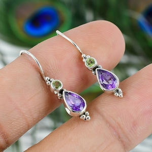 May include: A pair of silver dangle earrings with a teardrop-shaped amethyst gemstone and a smaller round peridot gemstone. The earrings have a delicate design with small silver beads.