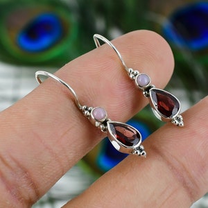 May include: A pair of silver dangle earrings featuring a teardrop-shaped red gemstone and a small pink gemstone on each earring.