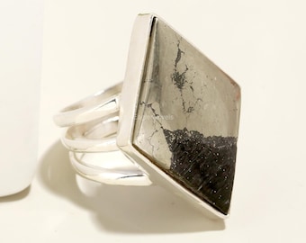 Puede incluir: Un anillo de plata con una piedra preciosa de pirita de forma cuadrada. El anillo tiene una banda ancha con una superficie texturizada.