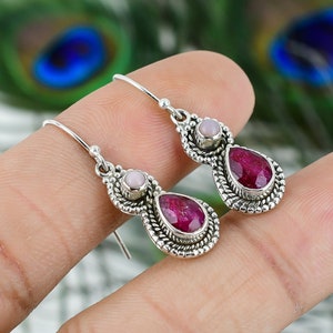 May include: Silver dangle earrings featuring a teardrop-shaped, deep red gemstone and a smaller, round, light pink stone. The gemstones are set in an ornate, textured silver setting.