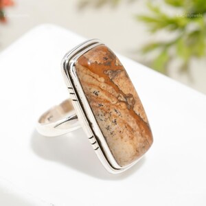 May include: A rectangular ring with a large, polished stone in tan and brown shades. The stone is set in a silver band with a simple design. This statement ring is a striking piece of jewellery.