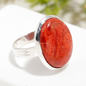 May include: A silver ring featuring a large, oval-shaped red coral stone. The stone has a natural, textured surface.